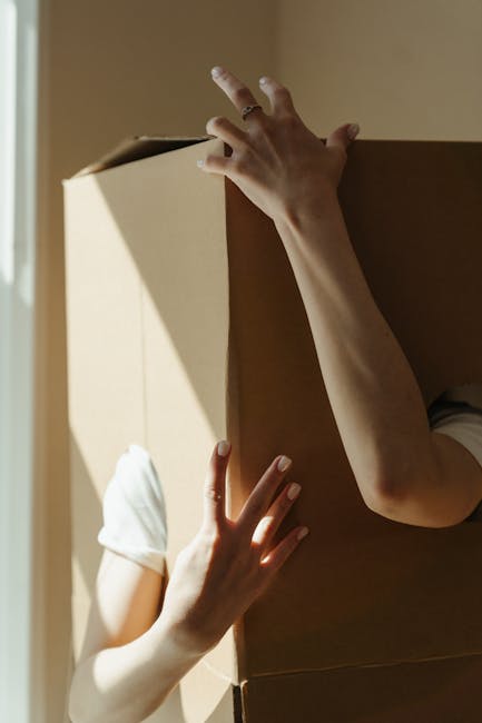 A person wearing a white t-shirt and blue jeans is kneeling indoors on a light-colored floor, writing on a cardboard box with a red marker. The box is sealed with white packing tape across the top. In the background, there are additional cardboard boxes of various sizes, some open and some closed, indicating packing and moving preparations. The scene appears to take place in a home environment, possibly a hallway or storage area, with natural lighting illuminating the space. The individual is in the process of organising items for home relocation or furniture transport, which is part of a professional removal service operated by Man with Van New Addington. The overall setting reflects the packing and loading process typical of house removals and detailed moving logistics.