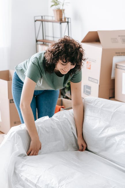 A woman with curly brown hair, dressed in a green t-shirt and blue jeans, is seen preparing a large white wrapped item, possibly furniture or household goods, on a sofa inside a residential room. Behind her, there are several cardboard boxes, some open and others sealed, indicating an ongoing packing process as part of house relocation or moving preparations. A metal shelving unit with potted plants and other household items is visible in the background, along with natural light coming through a window. The scene depicts the packing stage of a home move, involving careful wrapping and organizing of larger items for transportation, reflecting the services offered by Man with Van New Addington for efficient furniture transport and removals.