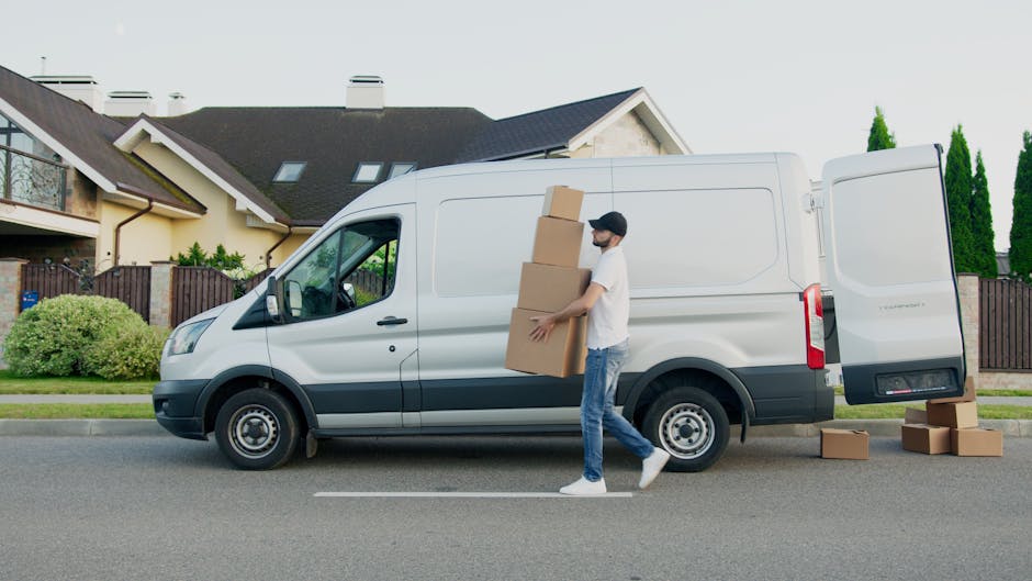 A man wearing a white t-shirt, jeans, and a baseball cap is walking alongside a white cargo van parked on the street in New Addington during daytime. He is carrying three cardboard boxes stacked verti