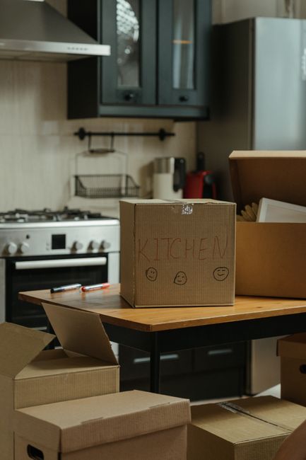Inside a home kitchen during a move, a cardboard box labeled 'KITCHEN' with handwritten smiley and neutral faces is placed on a wooden dining table. Surrounding the box are several other packed boxes made of corrugated cardboard and packed with packing paper or bubble wrap. The kitchen features dark-colored cabinets mounted on the wall above a white stove and oven, with a tiled backsplash. A metal shelf holds a wire rack, and various small appliances and utensils are visible on the counter. The scene captures a moment of packing and preparation for furniture transport or home relocation, with natural indoor lighting highlighting the organized packing process managed by Man with Van New Addington, a professional removals service specializing in house removals and moving logistics.