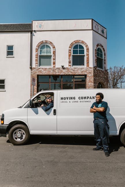 A white moving van parked on the street outside a modern, two-storey building with a brick and white facade, large front windows, and arched window frames. The van has the words 'MOVING COMPANY' and 'LOCAL & LONG DISTANCE' printed on its side. Inside the driver’s cabin, a person is visible, engaged in a conversation with a crew member standing outside the vehicle. The crew member, dressed in dark clothing, is leaning against the van with arms crossed, smiling, and appears ready for a home relocation or furniture transport. The scene is set in daylight with clear blue sky, and the environment suggests a residential or commercial area suitable for house removals or moving services, reflecting the activities involved in packing, loading, and transporting belongings for a house move in New Addington.