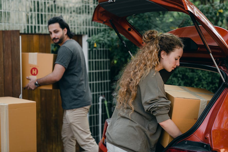 A woman with long, curly blonde hair, dressed in a gray jacket, is loading a cardboard box into the open trunk of a red hatchback vehicle parked outside a residential property. Inside the trunk, additional boxes are visible, indicating a packing and moving process. In the background, a man with dark hair and a beard, wearing a gray T-shirt and light-colored pants, is walking toward the property while carrying a cardboard box labeled with a red 'H'. The setting appears to be a backyard or garden with a wooden fence and green foliage, and the scene is lit with natural daylight. The image captures elements related to home relocation, including packing materials, furniture, and the equipment used for moving, aligning with professional removals services provided by Man with Van New Addington during furniture transport and loading activities.