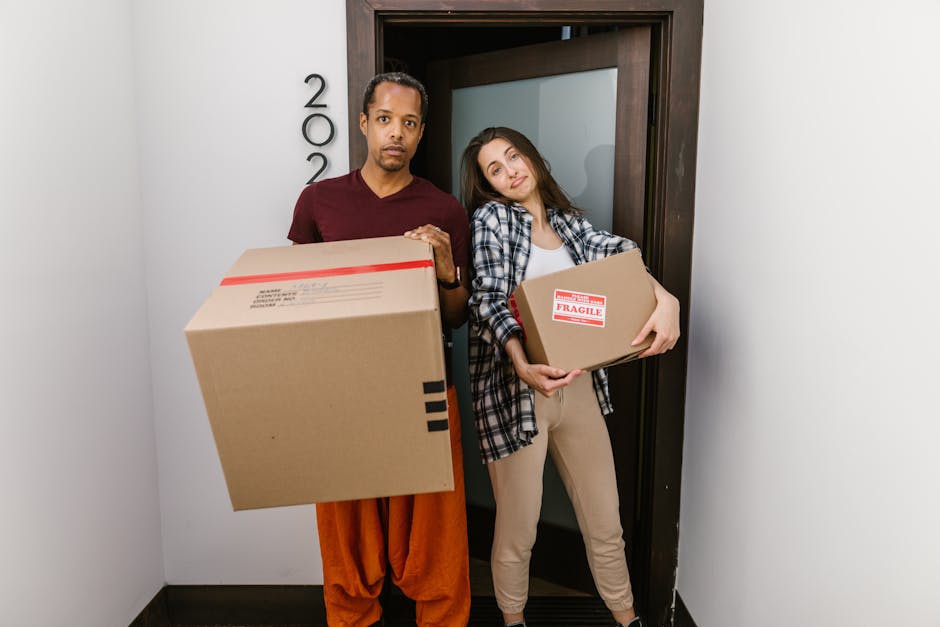 A man and a woman standing inside a hallway in front of a door numbered 200, both holding cardboard moving boxes during a home relocation process. The man, dressed in a maroon t-shirt and orange pants