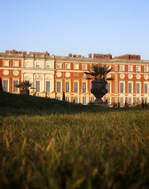 A large historic building with ornate architectural details and multiple windows, situated behind a well-maintained grassy area. In the foreground, a decorative stone or ceramic planter holding a palm tree is visible, with sunlight casting a warm glow on the facade. The image appears to be taken from a low angle, emphasizing the height of the building and the outdoor space. This scene is representative of the type of property that might be involved in home relocations or furniture transport services offered by Man with Van New Addington in the process of packing and moving near Addington Palace.