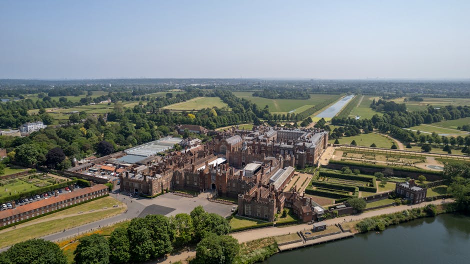 An aerial view of a large, historic castle estate surrounded by extensive landscaped gardens, green fields, and farmland. The castle features multiple towers, turrets, and ornate brickwork, with several adjoining buildings and a lengthy glass-roofed structure. The estate is bordered by a river or canal along one side, with pathways and roadways connecting different sections. Nearby, a parking area is visible with several vehicles, and a road runs adjacent to the property. The surrounding landscape is lush and verdant, with trees and open fields, under a partly cloudy sky. This image exemplifies the type of property that may be involved in home relocation or furniture transport within house removals services, as provided by Man with Van New Addington.