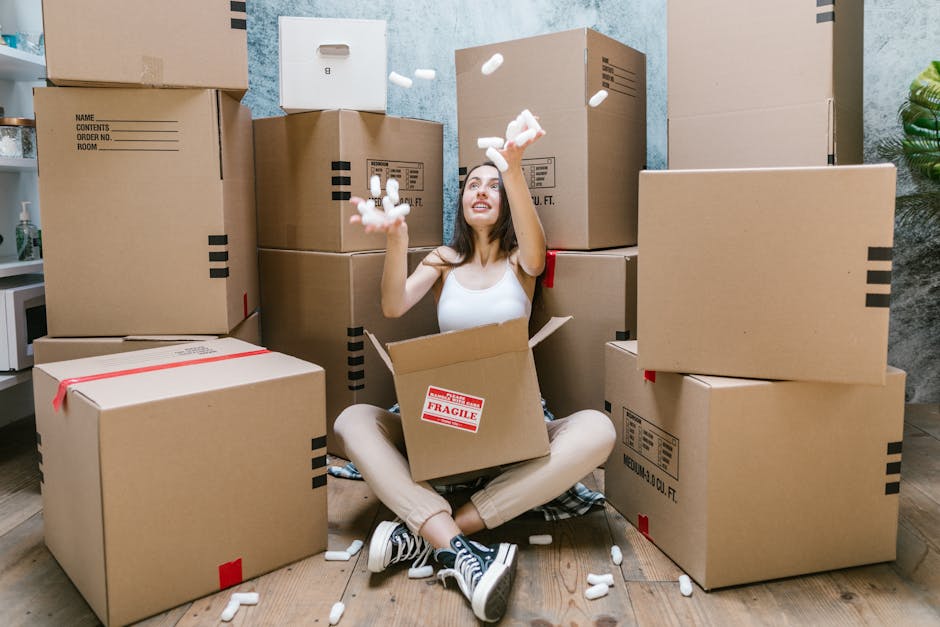A young woman sitting cross-legged on a wooden floor inside a home, surrounded by large cardboard moving boxes, with some boxes open and packing foam packaging scattered around her. She is wearing a white tank top, beige pants, and striped sneakers, and appears to be in the process of packing or unpacking. The background shows a partially visible kitchen area with a microwave and some household items, alongside a textured blue wall. The scene depicts a home relocation or packing process associated with professional removals services, such as those provided by Man with Van New Addington, illustrating the typical logistics involved in furniture transport and home moving activities.
