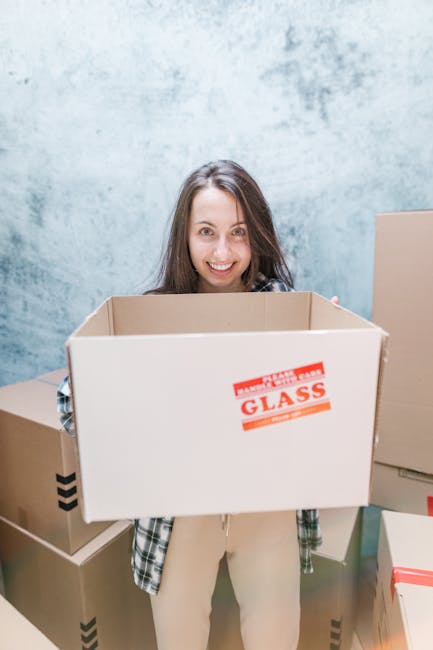 A woman with long brown hair smiling and standing indoors against a light blue textured wall, holding an open cardboard box labeled 'Fragile Glass' in red and orange print. She is surrounded by several other closed cardboard boxes of varying sizes, some with black arrow symbols indicating orientation, suggesting an ongoing home relocation or packing process. The boxes appear to be made of sturdy brown kraft cardboard, with some wrapped in plastic or tape, and are positioned on the floor and nearby surfaces. The lighting is bright and even, highlighting the neat stacking and organization typical of professional removals services. This scene reflects the packaging and loading stage of furniture transport and packing during a house removal, consistent with the type of service offered by Man with Van New Addington.