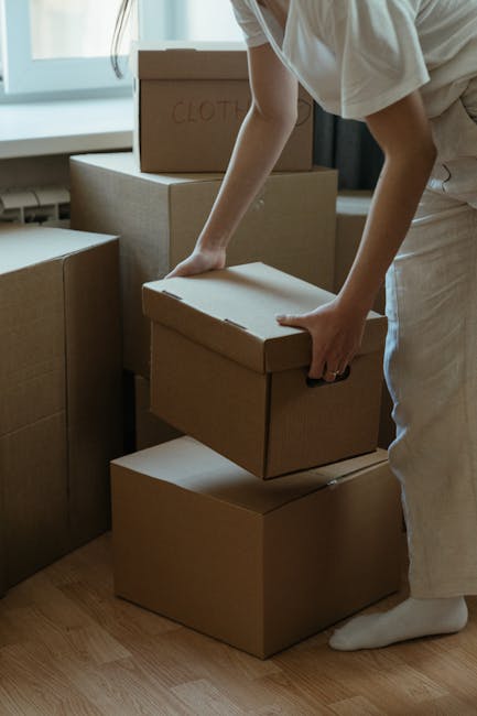 A young woman sitting cross-legged on a wooden floor inside a home, surrounded by large cardboard moving boxes, with some boxes open and packing foam packaging scattered around her. She is wearing a white tank top, beige pants, and striped sneakers, and appears to be in the process of packing or unpacking. The background shows a partially visible kitchen area with a microwave and some household items, alongside a textured blue wall. The scene depicts a home relocation or packing process associated with professional removals services, such as those provided by Man with Van New Addington, illustrating the typical logistics involved in furniture transport and home moving activities.