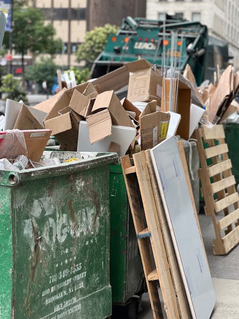 A large green waste disposal bin filled with various packing materials and small cardboard boxes, positioned on a street beside a wooden pallet with a flat-screen television and packaging foam. Behind the bin, a waste collection vehicle is visible, indicating a municipal or commercial rubbish collection area. The scene includes a background of urban buildings and trees, with natural daylight illuminating the area. This image reflects the disposal of packing waste and unwanted items, relevant to house removals and packing processes handled by Man with Van New Addington, a professional moving company. The setting emphasizes the importance of proper waste management during home relocation and moving logistics, supporting the themes of packing, furniture transport, and loading in the context of removals and disposal regulations in New Addington.