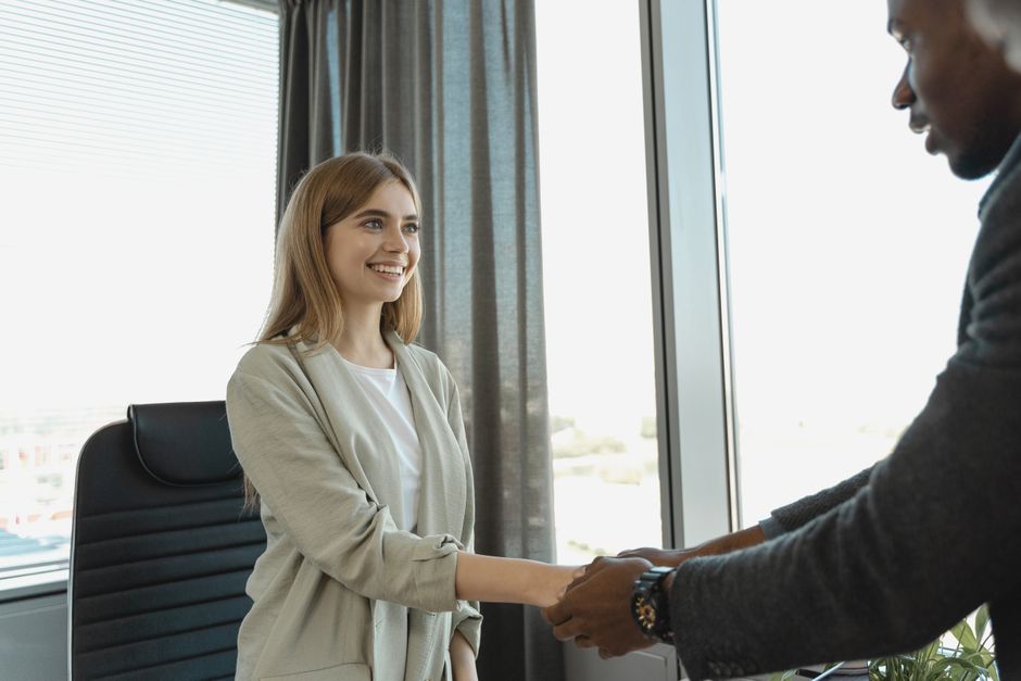 A professional moving assistant from Man with Van New Addington greets a young woman inside an office during a home relocation process. The assistant, dressed in formal attire, is extending his hand for a handshake, while the woman, with long blonde hair and a light-colored blazer, smiling warmly, responds to the greeting. The room has a large window with grey curtains, allowing natural daylight to illuminate the scene, and a black office chair is visible behind the woman. This interaction likely marks the beginning of a furniture transport or packing and moving service, with the assistant prepared to assist with moving logistics, including loading plans or initial consultations for household or office removals. The setting emphasizes professionalism and smooth client engagement during the house removal process, aligning with the content about skip hire rules and council disposal guidance in New Addington.