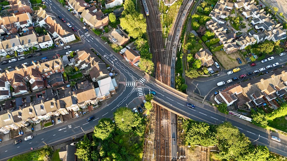 An aerial view of a residential neighbourhood showing rows of terraced houses with pitched roofs, surrounded by green trees and gardens. The image captures a busy intersection where multiple roads converge, with vehicles including cars and vans visible on the streets. A railway bridge crosses over the roads, with railway tracks running through the middle of the scene, supported by metal structures. The surrounding streets are lined with parked cars, and the area appears to be in daylight with clear visibility of the environment. This scene illustrates the typical urban setting where house removals and home relocation services provided by Man with Van New Addington may operate, highlighting the importance of efficient transport logistics in moving furniture and packed boxes through complex urban infrastructure.