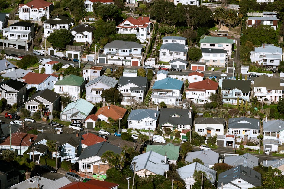 Aerial view of a residential neighbourhood in New Addington, showing a roundabout with multiple lanes and small traffic islands at the intersection. Surrounding the roundabout are closely packed houses with brick or tiled roofs, some with visible gardens enclosed by fences or hedges. Several parked cars line the streets, with a variety of vehicle types including small cars and vans. Green trees and lush vegetation are seen throughout the area, providing a contrast to the urban landscape. In the foreground, a building with a flat roof appears to be a commercial or community premises, featuring a parking lot with arranged parking spaces filled with cars. The overall scene depicts a typical suburban area with calm traffic flow, reflecting aspects of home relocation and furniture transport processes supported by professional removal services like those offered by Man with Van New Addington. The lighting indicates daytime, with natural light illuminating the scene clearly.